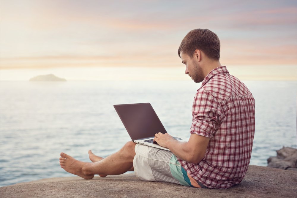 man with laptop sitting next to ocean while doing remote jobs online