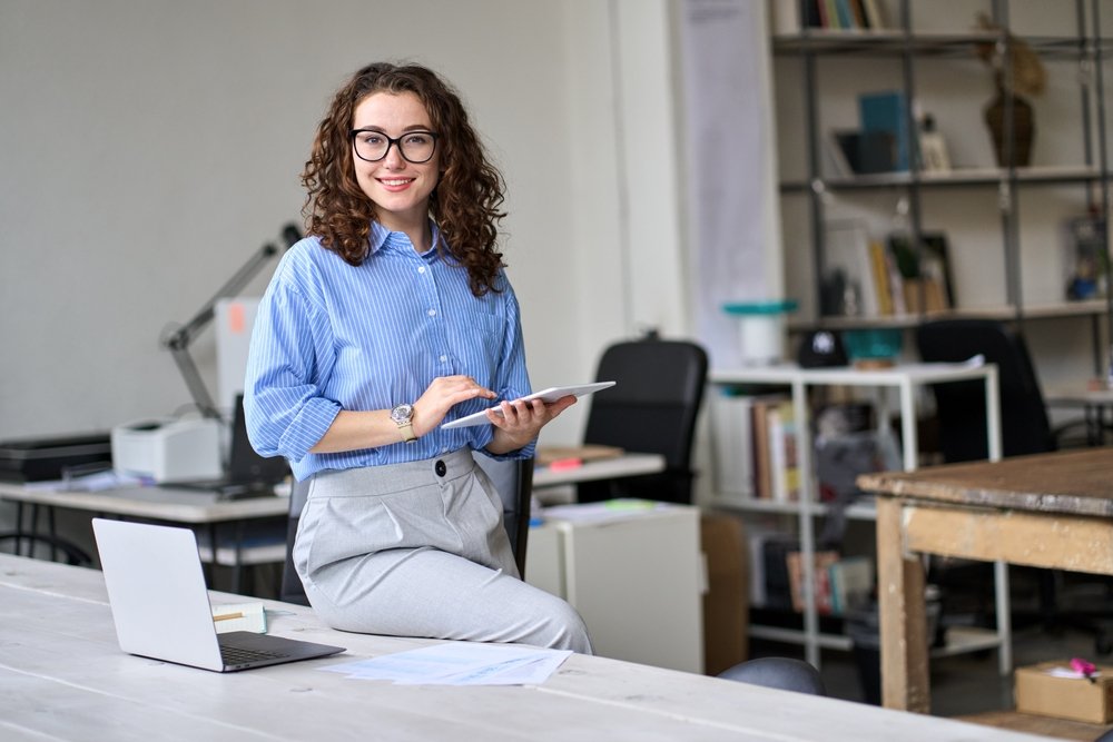 A female sales rep that has been recruited for remote sales jobs and she is sitting on the desk with her laptop and ipad on her hand