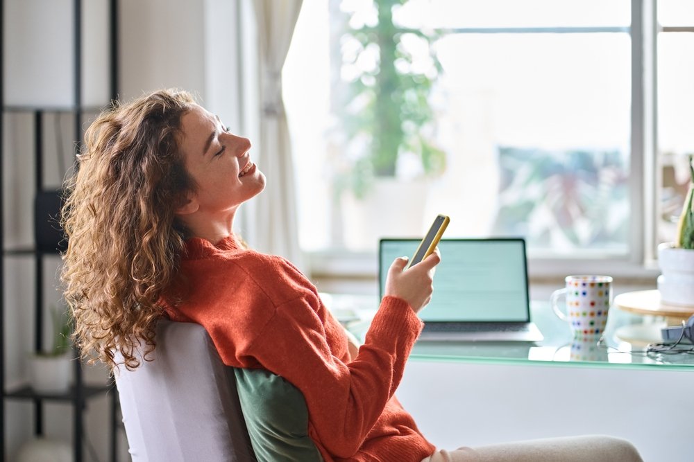 Remote Work Life: young smiling pretty woman holding smartphone while taking a break from working working on her laptop