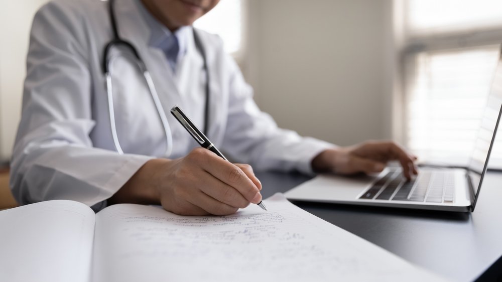 Remote Medical Coding Jobs: A picture showing a woman doctor in white uniform with stethoscope taking notes, using laptop, writing in medical journal, professional therapist practitioner filing documents or patient card