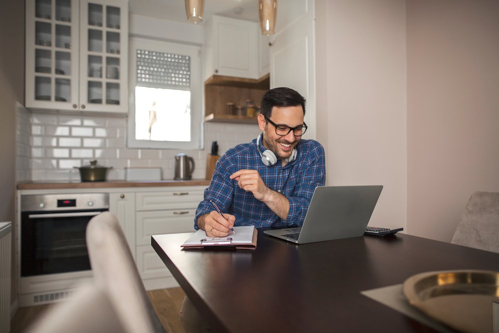 Overnight Remote Jobs: Young smiling man working from home writing notes in notebook while using his laptop and wearing headphones around his neck.