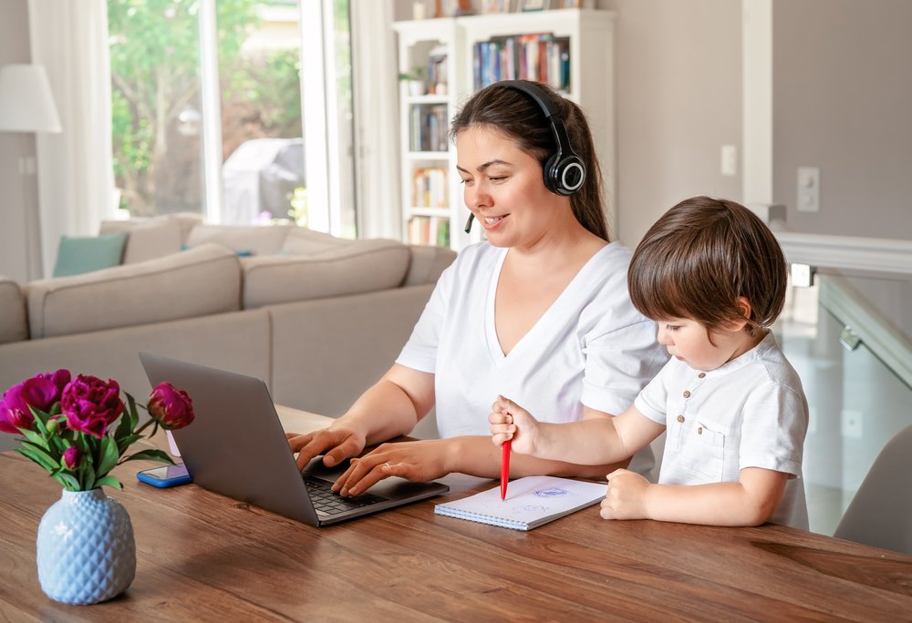 The Best and Worst Aspects of Remote Work: Freelance, online work, studying, learning with child concept. Home office. Woman with headphones working remotely on laptop while her little son drawing next to her. 