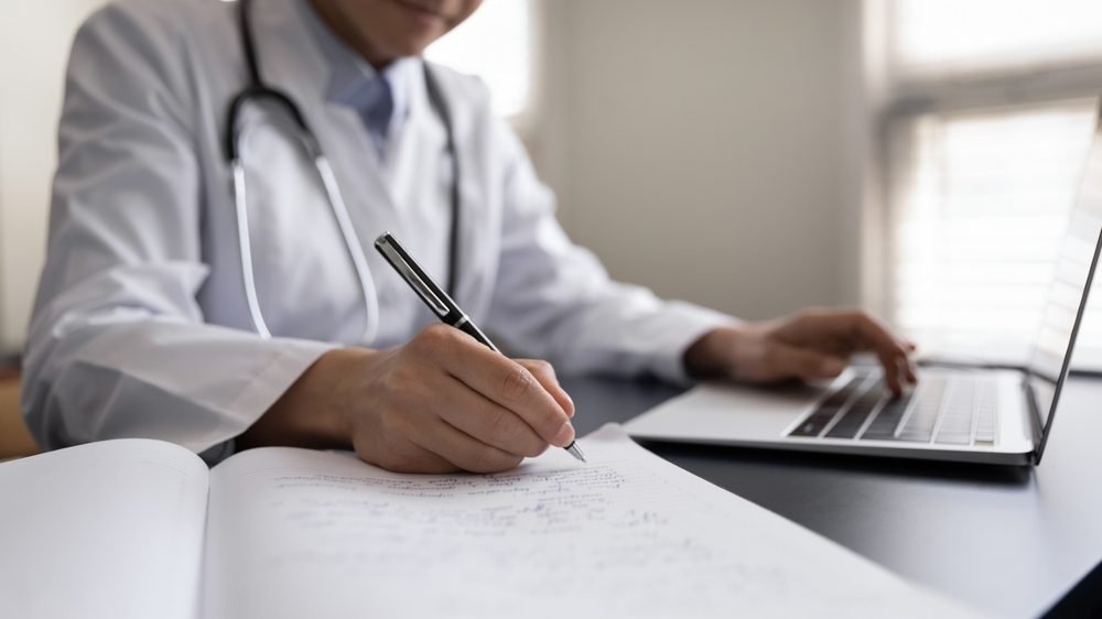 Remote Medical Coding Jobs: A picture showing a woman doctor in white uniform with stethoscope taking notes, using laptop, writing in medical journal, professional therapist practitioner filing documents or patient card