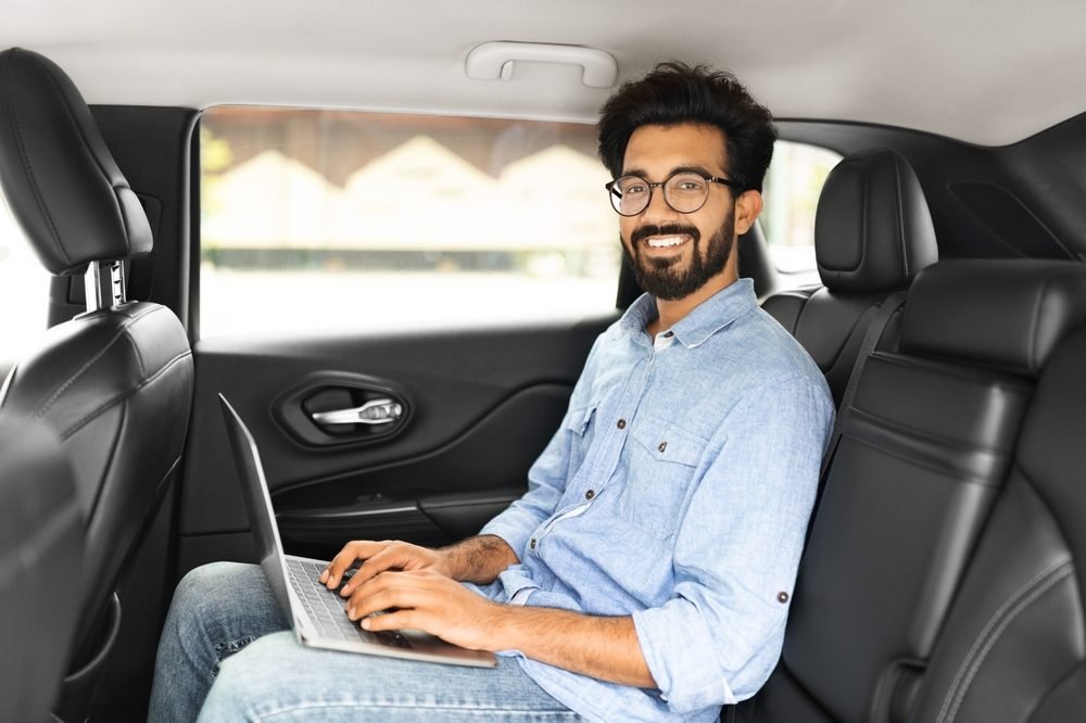 Top 5 Secrets to Boost Your Productivity While Working From Home: Cheerful handsome millennial eastern guy in casual wearing eyeglasses sitting on auto back seat, using laptop and smiling, man working remotely online while sitting in taxi