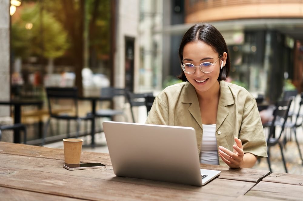 Full-Time vs. Part-Time Remote Jobs: Young woman sitting on online meeting in outdoor cafe, talking to laptop camera, explaining something, drinking coffee.