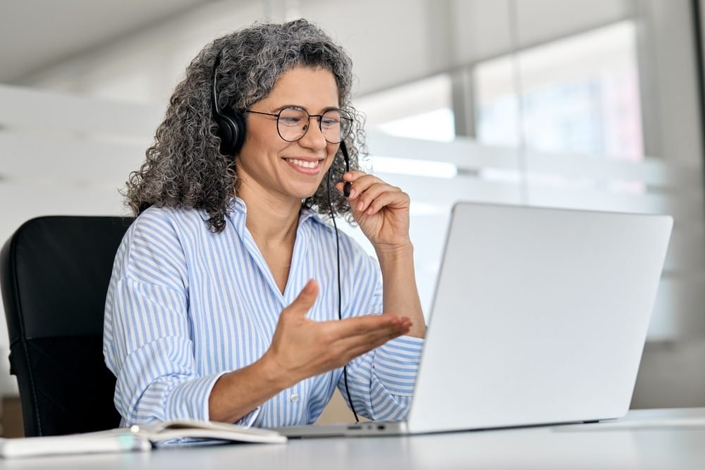Remote Customer Service Jobs: Happy mature business woman, a call center representative, a customer support agent talking to a client, smiling middle aged senior female operator wearing a headset working using a laptop computer in the office.