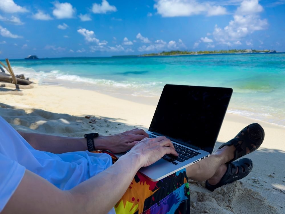 Working Normads: Nomad digital young man with the laptop and running remotely with bright scenic view near poolside on the beach in summer time